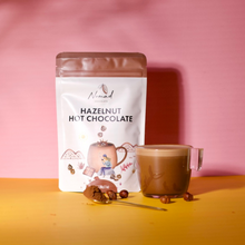 Load image into Gallery viewer, Image of Nomad chocolate hazelnut 200g with hazelnuts, spoon with cacao powder and a glass cup with hot chocolate drink.