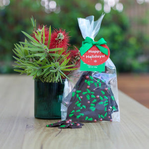 Image of chocolate bark in a cellophane bag with green ribbon and the swing tag saying happy holidays, next to it is a cup with a Australian botanical all sitting on a table