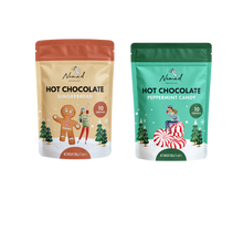 Load image into Gallery viewer, Two stand-up pouches of Nomad Visions hot chocolate mix shown side by side on a white background. The left pouch is gingerbread with a warm beige/tan background and orange-red top band, featuring a gingerbread man illustration.