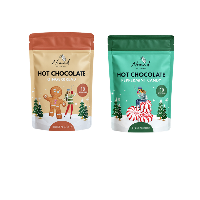 Two stand-up pouches of Nomad Visions hot chocolate mix shown side by side on a white background. The left pouch is gingerbread with a warm beige/tan background and orange-red top band, featuring a gingerbread man illustration.