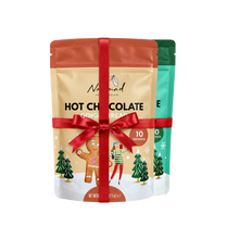 Load image into Gallery viewer, Two stand-up pouches of Nourish Chocolate Hot Chocolate Gingerbread flavor are shown together on a white background, wrapped with a red ribbon. The front pouch is beige with gingerbread-themed illustrations and a red ribbon bow, while the rear pouch is teal-green.