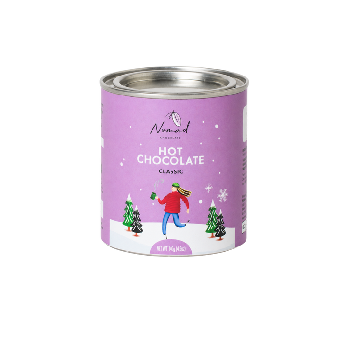 A 4.5 oz can of Nomad Chocolates Hot Chocolate Classic on a solid white background. The can features a light purple label with an illustration of a person running in a snowy landscape with Christmas trees.