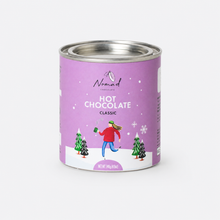 Load image into Gallery viewer, A can of Nomad Chocolate Hot Chocolate Classic on a solid white background. The can features a light purple label with an illustration of a person in a red jacket and blue pants running through a snowy landscape with Christmas trees.