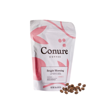 Load image into Gallery viewer, Minimalist product shot of Conure Coffee’s Bright Morning and Original Blend bags displayed on a light background with a ceramic cup of black coffee. Soft pastel tones and clean design highlight the premium, small-batch roasted coffee by Nomad Chocolate