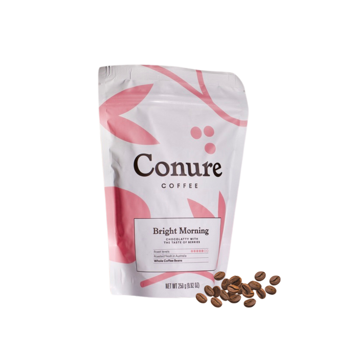 Minimalist product shot of Conure Coffee’s Bright Morning and Original Blend bags displayed on a light background with a ceramic cup of black coffee. Soft pastel tones and clean design highlight the premium, small-batch roasted coffee by Nomad Chocolate