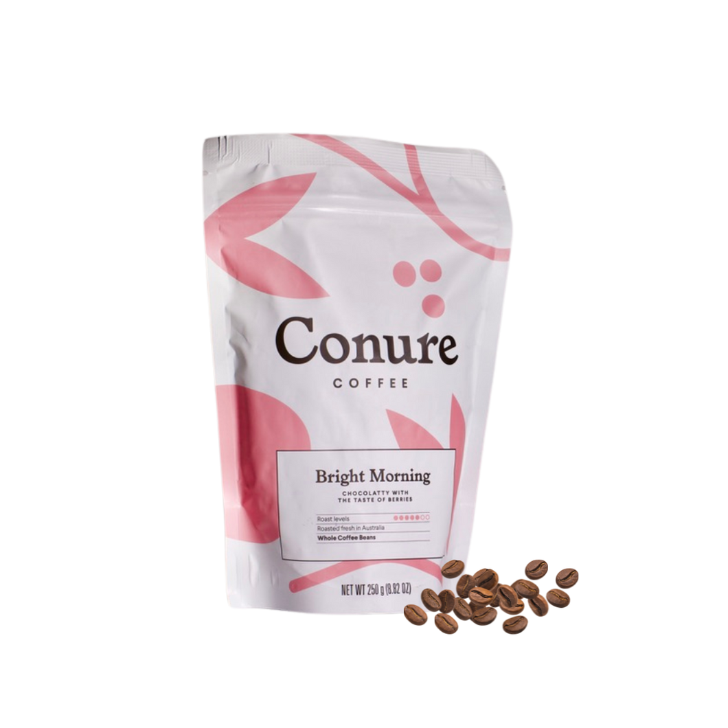 Minimalist product shot of Conure Coffee’s Bright Morning and Original Blend bags displayed on a light background with a ceramic cup of black coffee. Soft pastel tones and clean design highlight the premium, small-batch roasted coffee by Nomad Chocolate
