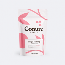 Load image into Gallery viewer, Conure Coffee Bright Morning chocolate coffee beans in a white stand-up pouch with pink leaf graphics and black branding, photographed on a light gray background.