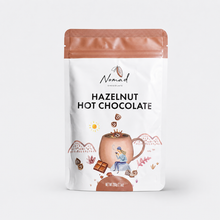 Load image into Gallery viewer, A stand-up pouch of Nomad Chocolate Hazelnut Hot Chocolate on a light gray background. The pouch is white with a metallic copper-rose top and bottom, featuring the brand name 'Nomad Chocolate' and the product name 'HAZELNUT HOT CHOCOLATE' in black text.