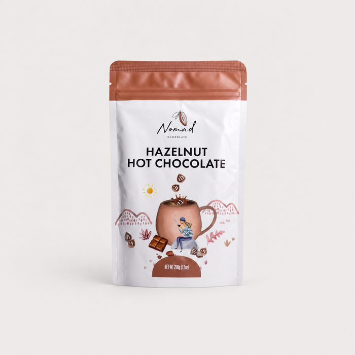 A 300g stand-up pouch of Nomad Chocolates Hazelnut Hot Chocolate, featuring a white bag with a copper-brown top, a central illustration of a hot chocolate mug, and black text on a clean, light off-white background.