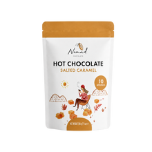 Load image into Gallery viewer, A stand-up pouch of Nomad Chocolate Hot Chocolate Salted Caramel, featuring a white body with an orange top and a whimsical illustration of a woman sitting by a hot chocolate cup near caramel cubes.