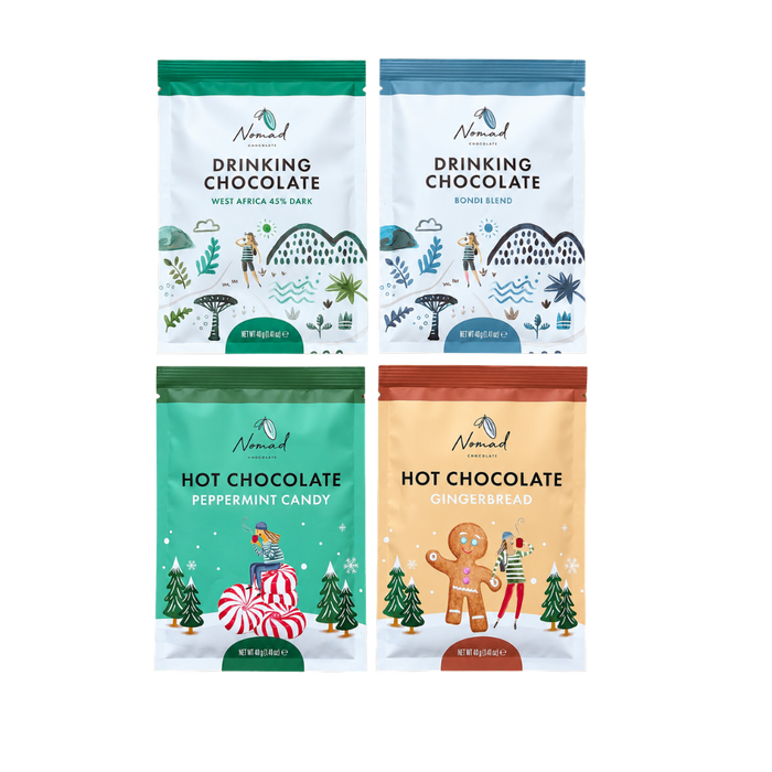 Four stand-up pouches of Nomad hot chocolates 40g arranged in a 2x2 grid on a plain white background. The top two are Drinking Chocolate bags with illustrated winter scenes in green and blue; the bottom two are Hot Chocolate bags with illustrated holiday scenes in teal and beige.