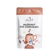 Load image into Gallery viewer, A stand-up pouch of Nomad Chocolate Hazelnut Hot Chocolate on a plain white background. The pouch is white with a copper-brown top seal and a central illustration of a mug, chocolate pieces, and decorative accents.