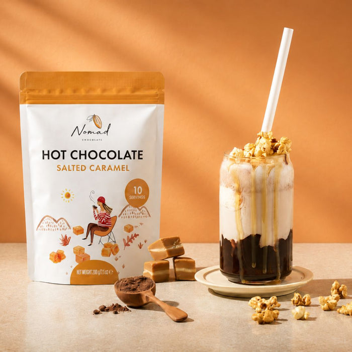Nomad Hot Chocolate Salted Caramel flavor pouch shown next to a caramel-flavored iced drink topped with caramel drizzle, popcorn, and a white straw, on a warm orange background.