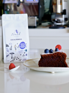 Lifestyle photo of chocolate cake slice dusted with icing sugar and decorated with fresh blueberries and raspberries. Nomad Chocolate Chef's Cocoa Powder Dominican 100% Dark, white packaging front. Girl wandering through the Nomad Chocolate landscape mixing a chocolate batter in a mixing bowl. Vegan, Gluten Free, Dairy Free, No Sugar