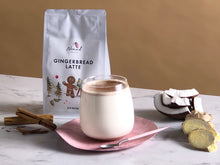 Load image into Gallery viewer, Nomad Chocolate Gingerbread Latte lifestyle picture with made latte dusted with cinnamon, cinnamon quills, coconut pieces and ginger slices on the side, white product bag. All natural ingredients, gluten free