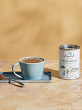 Load image into Gallery viewer, Nomad Chocolate West Africa 45% Dark white tin next to serving plate with hot chocolate in a green cup and spoon filled with cacao powder. 