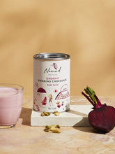 Load image into Gallery viewer, Nomad Chocolate Ruby Bliss organic with Beetroot and spices, glass with pink hot chocolate and halved fresh beetroot next to tin, sprinkled cardamon pods in front of tin