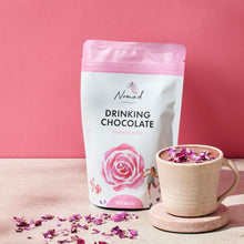 Load image into Gallery viewer, Nomad Drinking Chocolate Damask Rose bag next to cup with hot chocolate, topped with pink rose petals. Rose petals sprinkled in front of bag and cup.