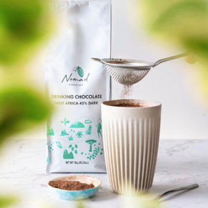Nomad Drinking Chocolate West Africa 45% Dark 1kg bag, lifestyle photo with hot chocolate in a cup and chocolate powder going through a sifter to dust the hot chocolate