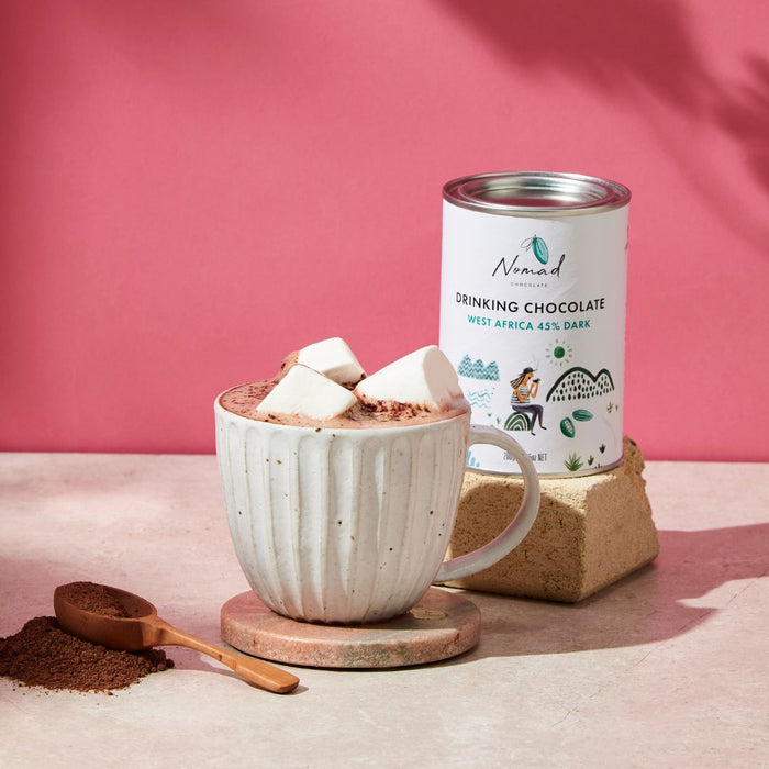 Nomad Chocolate West Africa tin next to mug with hot chocolate topped with white marshmallows and wooden spoon with cocoa powder next to cup.